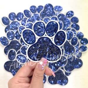May include: A navy blue sequined paw print patch with a white border, held in front of a background of similar paw prints. The patch is filled with small, shiny sequins. The overall theme is pet-related.
