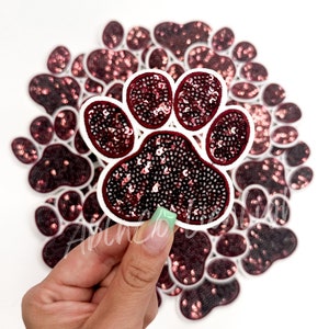 May include: A close-up of a burgundy sequined paw print iron-on patch. The patch is surrounded by other paw print patches.