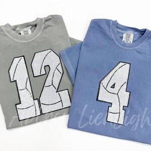 K&ouml;nnte beinhalten: Zwei T-Shirts mit Paillettenzahlen und einem Volleyball-Design. Das Shirt links ist grau mit der Nummer 12. Das Shirt rechts ist blau mit der Nummer 4.