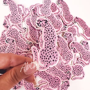 May include: A pile of pink cheetah iron-on patches. The patches are embroidered with black spots and have a white outline.