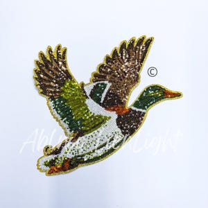 May include: A green, brown, and white sequined duck applique with gold trim. The duck is flying with its wings spread out.