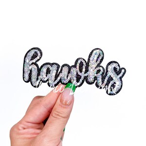 May include: A black glitter patch with the word "Hawks" in silver sequins. The patch is held against a white background. The patch is a decorative item.