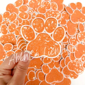May include: Orange paw print patches with white trim and sequin details. A hand holds one patch over a pile of similar patches. The patches are likely for crafts or embellishments.