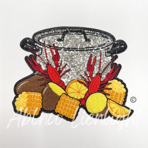 May include: A silver pot with a lid, surrounded by red crayfish, corn on the cob, potatoes, and lemons. The pot is covered in sequins.