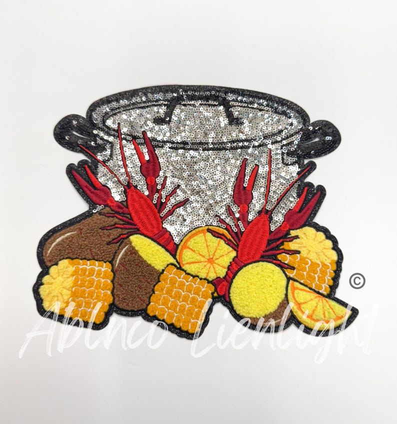 May include: A silver pot with a lid, surrounded by red crayfish, corn on the cob, potatoes, and lemons. The pot is covered in sequins.