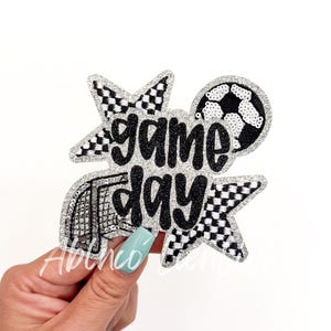 May include: A silver glitter iron-on patch with a black and white checkered star, a soccer ball, and the text "game day".