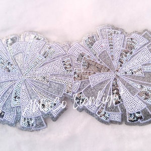 May include: Two silver sequin pom poms with white stitching. The pom poms are made of sequins and have a white outline.