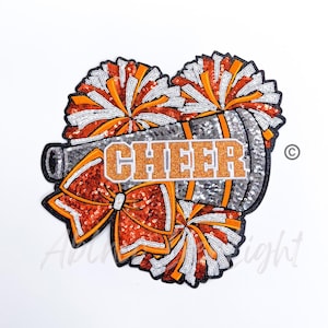 May include: A silver and orange sequin cheerleading patch with a megaphone, pom-poms, and a bow. The word "CHEER" is written in orange glitter on the megaphone.