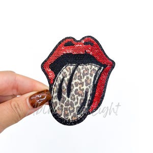 May include: A red sequined lips patch with a black outline and a leopard print tongue. The patch is held by a hand against a white background. The patch is a fun accessory for clothing or bags.