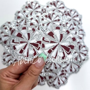 May include: A close-up of a pile of maroon and silver glitter cheer pom poms. The pom poms are made of fabric and have a white and silver glitter design.