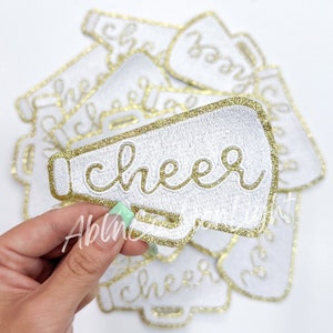 May include: White embroidered patch in the shape of a megaphone with the word "cheer" in gold script. The megaphone has a gold glitter border. Several similar patches are visible in the background.