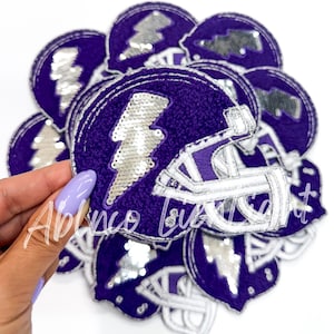 May include: Purple and silver iron-on patches featuring a football helmet with a lightning bolt design. The patches are made of a soft, fuzzy material and have a sparkly finish.