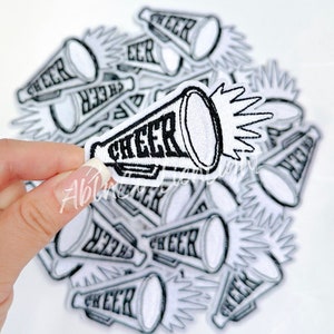 May include: A pile of white iron-on patches with a black outline of a megaphone with the word "CHEER" written on it.