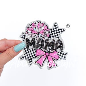 May include: A sequined patch with the word "MAMA" in black sequins. The patch features a pink bow, a pink pom-pom, and a checkered pattern. The edges are outlined in silver glitter.