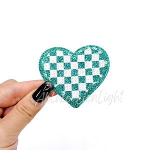 May include: A heart-shaped patch with a checkerboard pattern in white and teal. The patch is outlined with teal glitter. The patch is held by a hand with a black manicure. The text "Albinogreenlight" is partially visible.