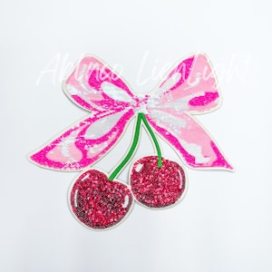 May include: Pink and white sequined bow with two red sequined cherries attached to the bow.