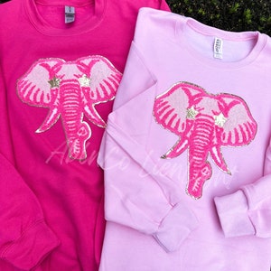 May include: Two pink sweatshirts with a pink glitter elephant design. The elephant has gold glitter stars on its head. One sweatshirt is a darker pink and the other is a lighter pink.
