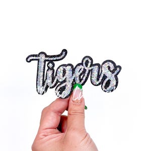 May include: A black fabric patch with the word "Tigers" in silver glitter lettering.