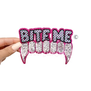 May include: A pink sequined patch with the words "BITE ME" in a checkered pattern of blue and black. Below the text are sequined fangs. The patch is held by a hand against a white background.