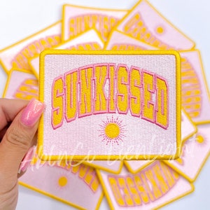 May include: A stack of pink and yellow embroidered iron-on patches with the word "SUNKISSED" in a retro font. The patches feature a yellow sun with pink rays.