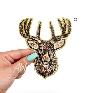 May include: A gold and brown sequined deer head applique patch. The deer has large antlers and a detailed face.