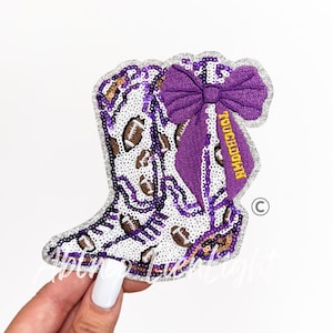 May include: A sequined patch in the shape of a cowboy boot, featuring footballs and a purple bow. The boot is white with brown footballs and purple accents. The bow has the word "Touchdown" on it.