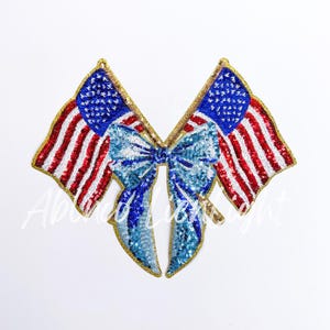 May include: A pair of American flag patches with a blue bow in the center. The patches are made of sequins and have a gold outline.