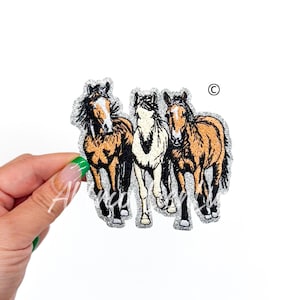 May include: An embroidered patch featuring three horses running side-by-side. The horses are brown, white, and brown with black markings. The patch is on a silver glitter background.