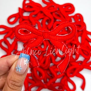 May include: A collection of bright red, embroidered bow appliques. One bow is held up, showcasing the intricate stitching and design. The bows are likely for crafting or embellishment. The text "Ablnco LienLight" is visible.