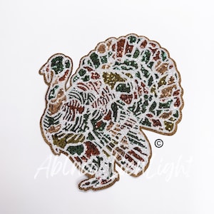 May include: A decorative turkey-shaped applique with a sequined design. The turkey features a brown outline and is filled with a mix of green, brown, and white sequins. The design is on a white background.