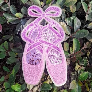May include: Pink ballet shoe shaped sequin patch with a bow. The patch is made of pink fabric and sequins.
