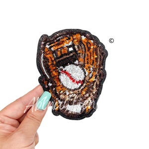 May include: A baseball glove and ball patch, embellished with sequins. The glove is brown and orange, with a white baseball featuring a red seam. The patch has a black border and is held by a hand with a light blue manicure.