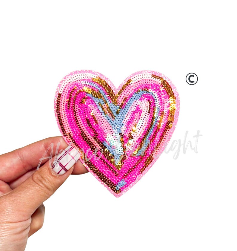 Puede incluir: Un parche de lentejuelas en forma de coraz&oacute;n en tonos rosa, dorado y azul claro. El parche se sostiene sobre un fondo blanco. Las lentejuelas est&aacute;n dispuestas en c&iacute;rculos conc&eacute;ntricos, creando un efecto texturizado. El parche es probablemente para fines decorativos.