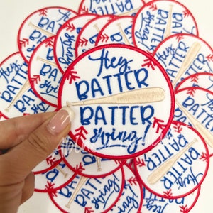 May include: A close-up of several embroidered iron-on patches with a baseball theme. The patches are white with red and blue stitching. The text on the patches reads "Hey Batter Batter Swing!"