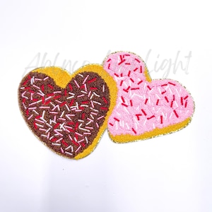 May include: Two heart-shaped cookies with pink and brown frosting and white and red sprinkles. The cookies are decorated with a gold outline.