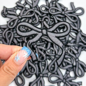 May include: A pile of gray fabric bow patches. One bow is being held up by a finger.