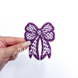 May include: Purple and white checkered bow with a glittery finish. The bow is a decorative embellishment for clothing or crafts.