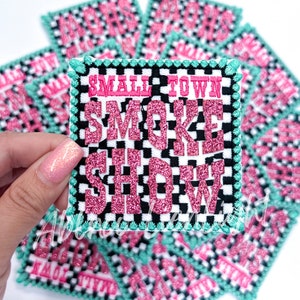 May include: Square patch with the text "SMALL TOWN SMOKE SHOW" in pink glitter lettering. The patch has a black and white checkered background, a teal border, and a white background. Several patches are visible.