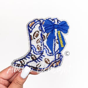 May include: A sequined patch in the shape of a cowboy boot, featuring footballs and blue accents. A blue bow is attached to the boot, with the word "TOUCHDOWN" in yellow. The patch is outlined in silver.