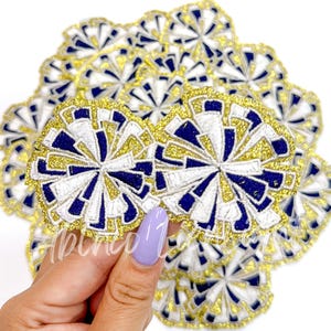 May include: A close-up of a stack of navy blue, white, and gold glitter iron-on patches. The patches are shaped like a flower with a circular design.