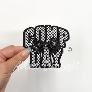 May include: Black and white checkered fabric patch with the word "COMP DAY" in black lettering and a black sequin bow in the center.