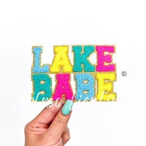 May include: A colorful felt applique with the words "LAKE BABE" in gold glitter letters. The letters are in different colors: blue, yellow, turquoise, and pink.