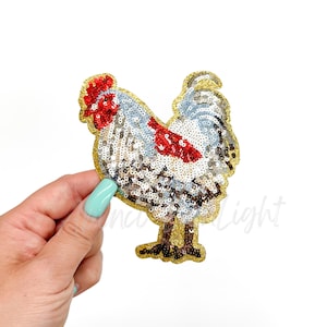 May include: A sequined rooster patch with a gold border. The rooster features red, white, and silver sequins, with a red comb and wattle. The patch is held by a hand with a light blue nail polish.