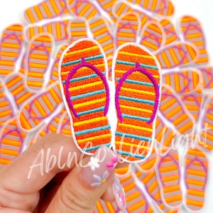May include: A close-up of two orange and blue striped flip-flop patches with pink straps. The patches are held in a hand with white flowers on the nails. There are many more of the same patches in the background.