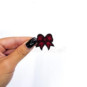 May include: A red fabric bow with black outline. The bow is small and is being held by a hand.