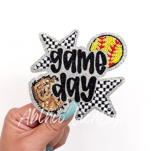 May include: A silver glitter iron-on patch with a black and white checkered star, a gold glitter softball, and the words "game day" in black.