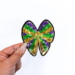May include: A green, purple, and gold sequined bow tie patch. The bow tie is made of sequins and has a black outline.