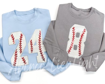 May include: Two gray and light blue sweatshirts with sequined baseball numbers. The gray sweatshirt has the number 8 and the light blue sweatshirt has the number 24.