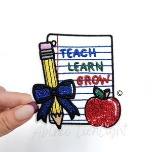 May include: An embroidered iron-on patch with a pencil, a red apple, and a piece of notebook paper with the words "Teach Learn Grow" written on it.