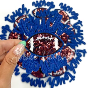 May include: A close-up of a pile of blue and white iron-on patches with the words "touch down" and a football design. The football is made of sequins and has a white and brown design.
